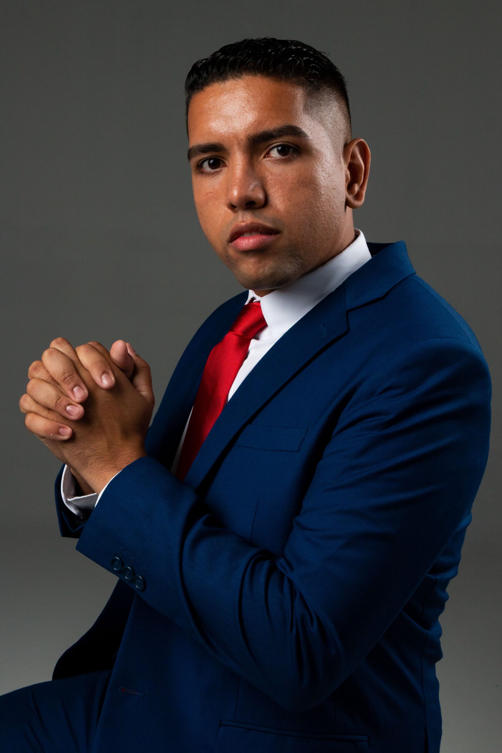 Professional studio portrait of a serious businessman in a blue suit with a red tie, hands clasped.